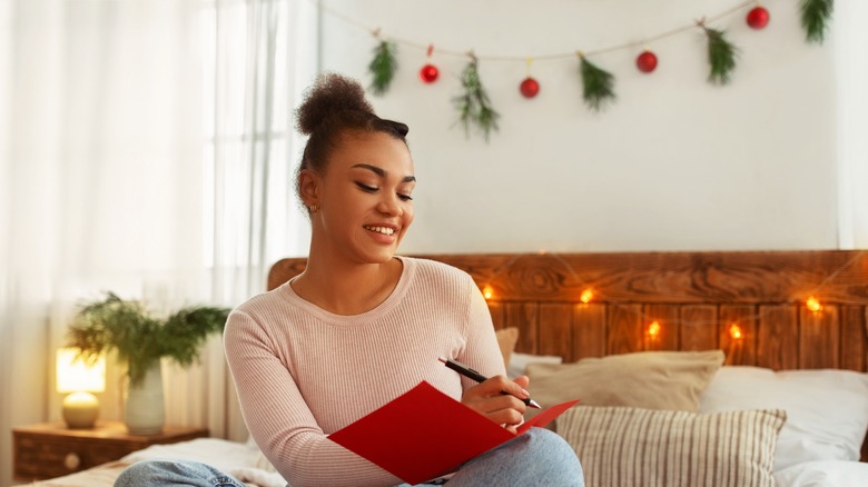 smiling young woman sitting on bed writing in red notebook with Christmas garland and lights in background