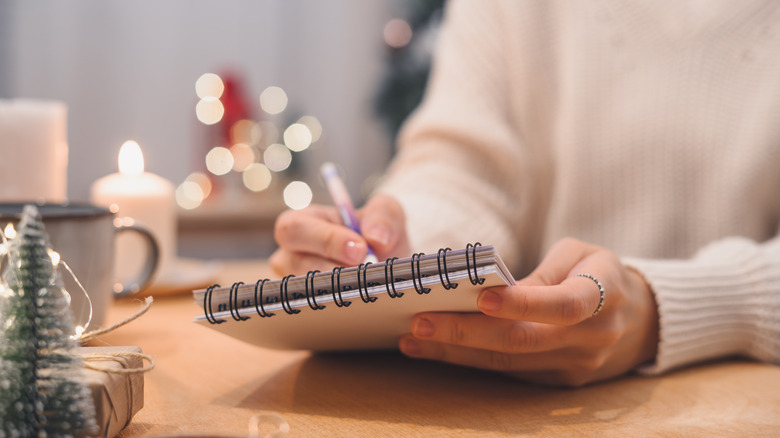 woman's hands writing in a spiral bound book with white Christmas lights and candles in background and mini bottle brush tree in foreground