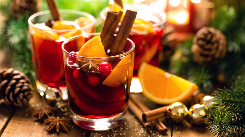 short glasses of mulled wine with star anise, cinnamon sticks, orange slices, and cranberries on wooden table with pine boughs, pine cones, and jingle bells