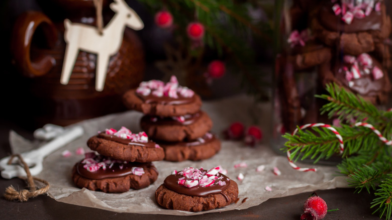 round chocolate cookies with chocolate ganache and crushed candy canes on parchment paper with pine branches and sugared cranberries around the side