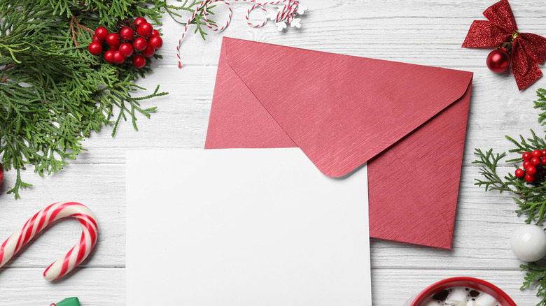 blank white invitation with red envelope on white wooden table with Christmas greens, red berries, ornament, and candy cane around sides