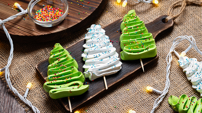 green and white meringue Christmas tree pops with round rainbow sprinkles on wooden cutting board and piece of burlap with white Christmas lights around them