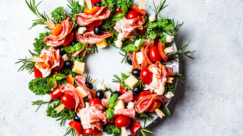 round plate with skewers of meat, olives, vegetables, and cheese arranged in shape of Christmas wreath with greens and herbs on white background