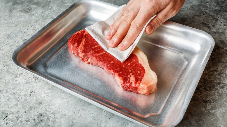 person patting raw steak dry with paper towel