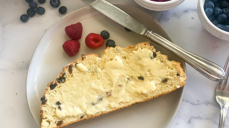 slice of buttered traditional Irish soda bread on a white plate with a knife near blueberries and red raspberries