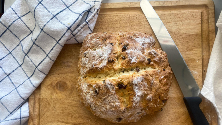 traditional Irish soda bread near a knife on a wooden cutting board near a blue and white checkered dish towel