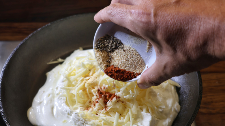 hand pouring small dish of spices into skillet with yogurt and shredded cheese