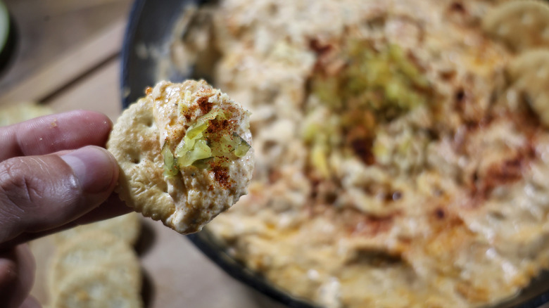 fingers holding cracker covered in Nashville hot chicken dip above skillet of the same