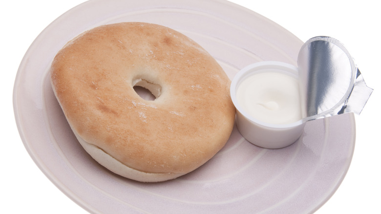 Bagel on a white plate with a little container of cream cheese