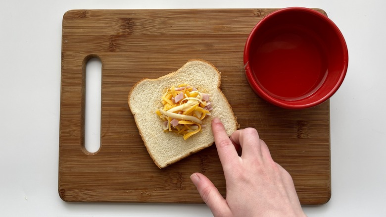 hand touching bread with jelly, chopped ham, and shredded cheese