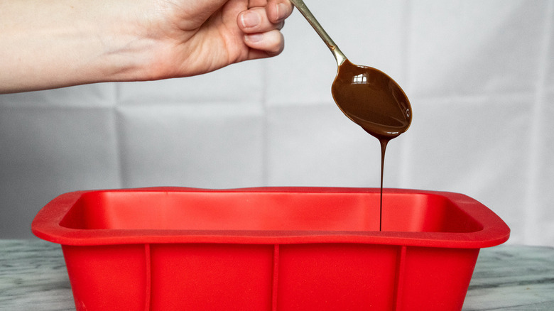 metal spoon drizzling chocolate onto the whipped cream layer in a red silicone loaf tin