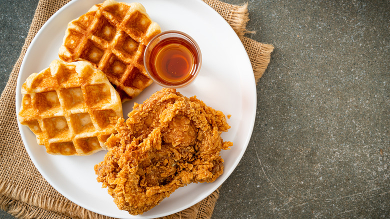 A plate of crispy fried chicken on a white plate with a jar of honey and waffles on a grey background