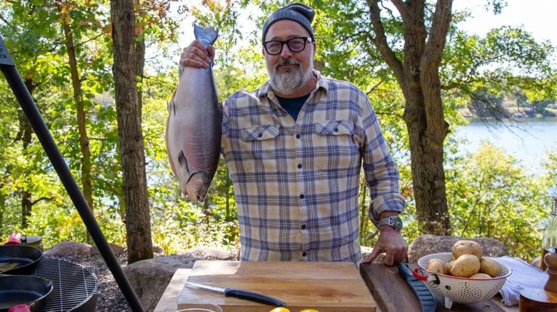 Andrew Zimmern in flannel shirt holding fish at outdoor cooking station