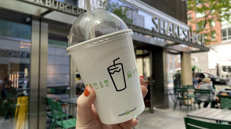 a hand holding a milkshake cup in front of a shake shack location