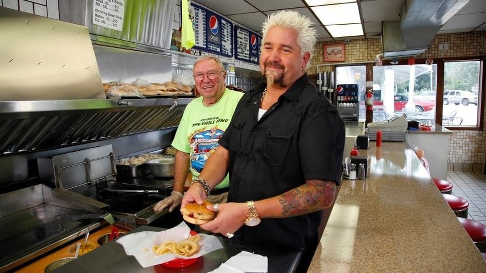 Guy Fieri smiles holding sandwich