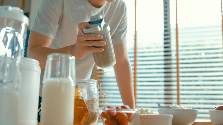 A man holding a shaker bottle of a protein shake with a blender and ingredients on a table in the foreground