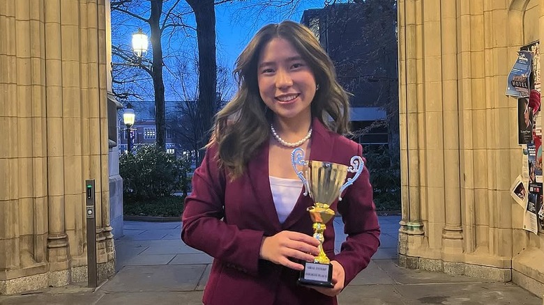 Smiling Liya Chu in maroon blazer holding trophy