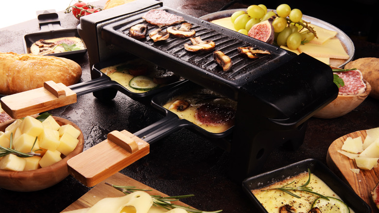 Raclette grill with mushrooms, melter plates, and plats of cheeses, fruits, and charcuterie