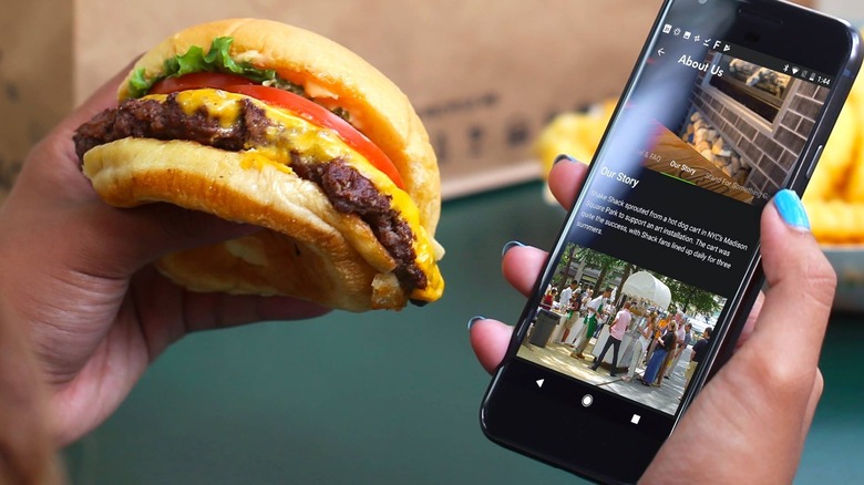 A left hand holding a Shackburger while the right hand has the Shack App open. Paper bag and fries in the background.