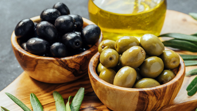 Green and black olives in olive wood bowls with olive oil in the background and olives leaves in the foreground