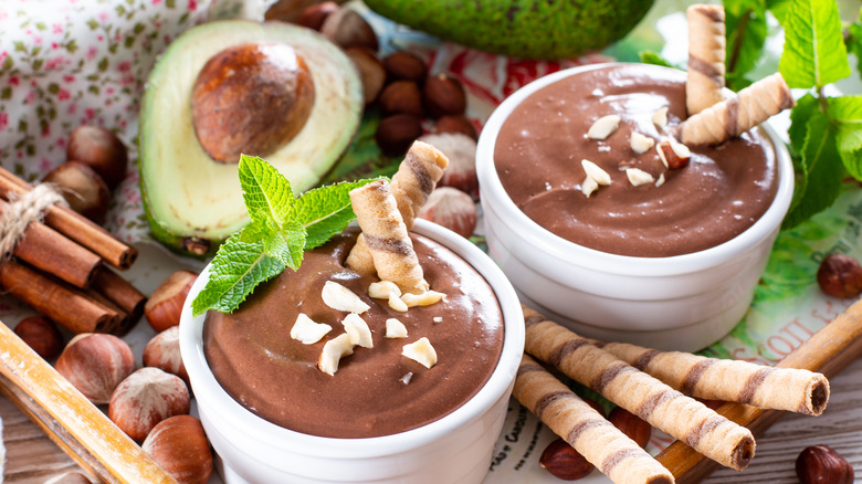 Raw avocado chocolate mousse in white cups garnished with cookies, nuts, and fresh mint with some nuts, cookies, and avocado on the side