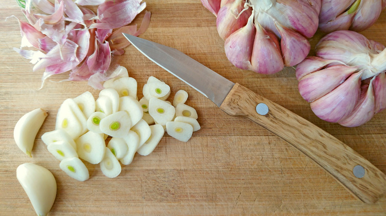 sliced fresh garlic on a cutting board with whole head of garlic, cloves, and a knife