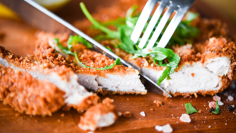 A fork cutting a crispy slice of pork covered with panko and topped with thinly sliced greens 