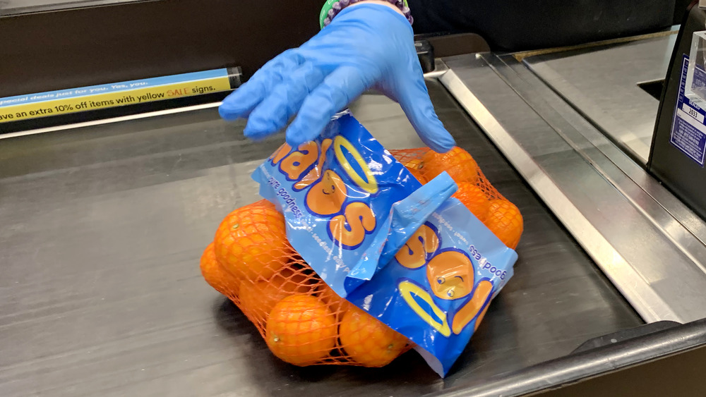 A gloved cashier dropping a pack of oranges onto the conveyor, conveying the contained nature of exploitative nature. of work in the pandemic... or something.