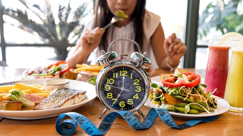 a women sits at a table surrounded by plates of food and a clock