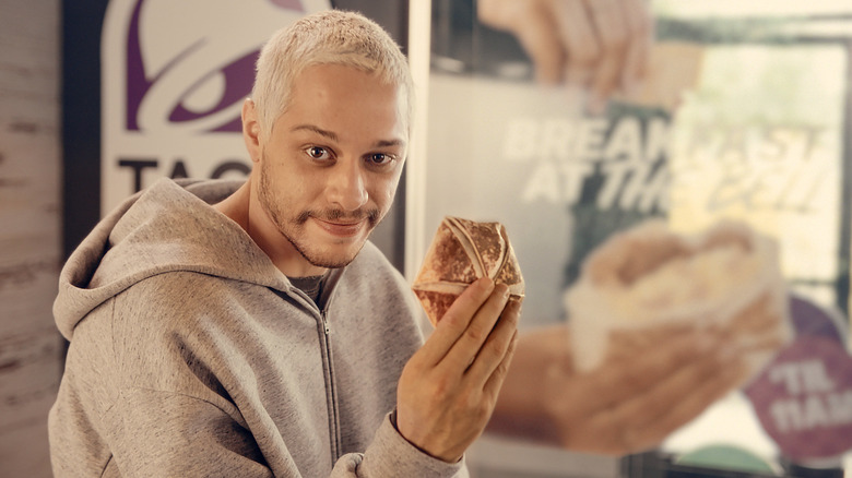 Pete Davidson holding a Taco Bell breakfast crunchwrap inside a Taco Bell restaurant