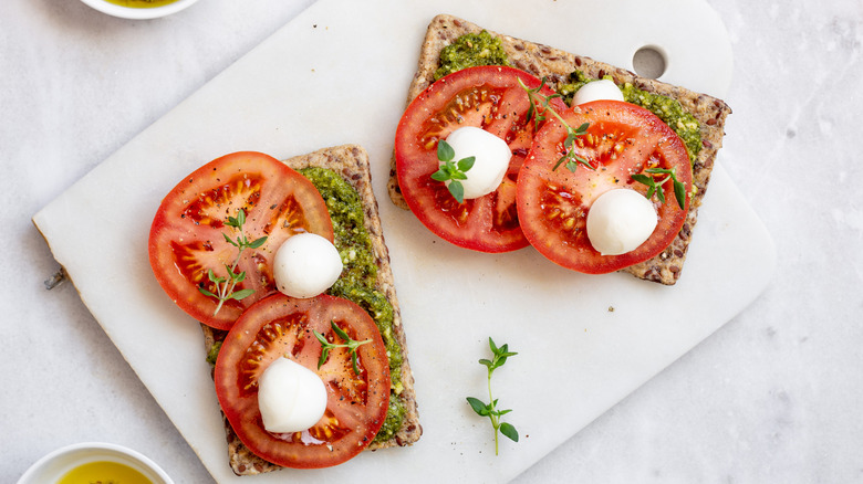 Two halves of a tomato, mozzarella, pesto sandwich on a cutting board