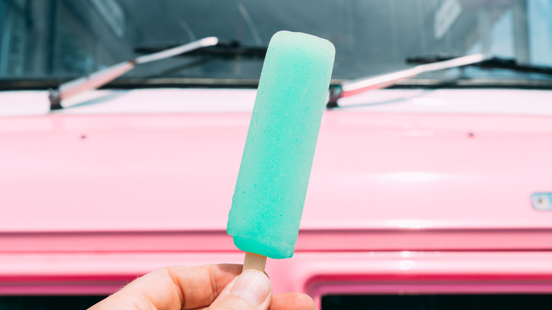 Hand holding a blue popsicle in front of an ice cream truck.