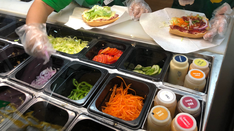 Two people making Subway sandwiches with containers of ingredients in the foreground. 