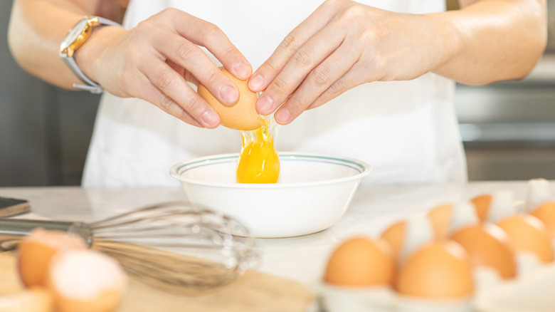 A person cracking an egg into a bowl surrounded by other eggs