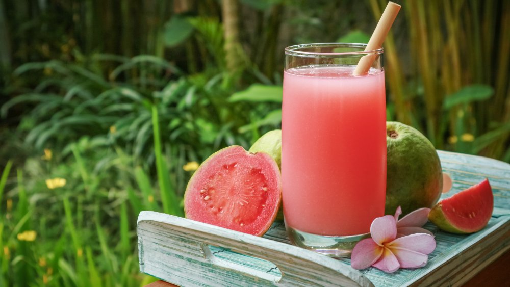 Glass of guava smoothie with a bamboo straw and cut guava on a wooden tray. There is a leafy outdoor background.