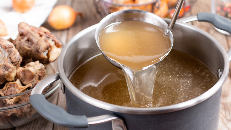 A ladle pouring broth into a pot with bones and onions in the background.