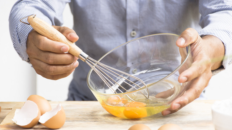 Man whisking eggs in a clear bowl with egg shells on a cutting board